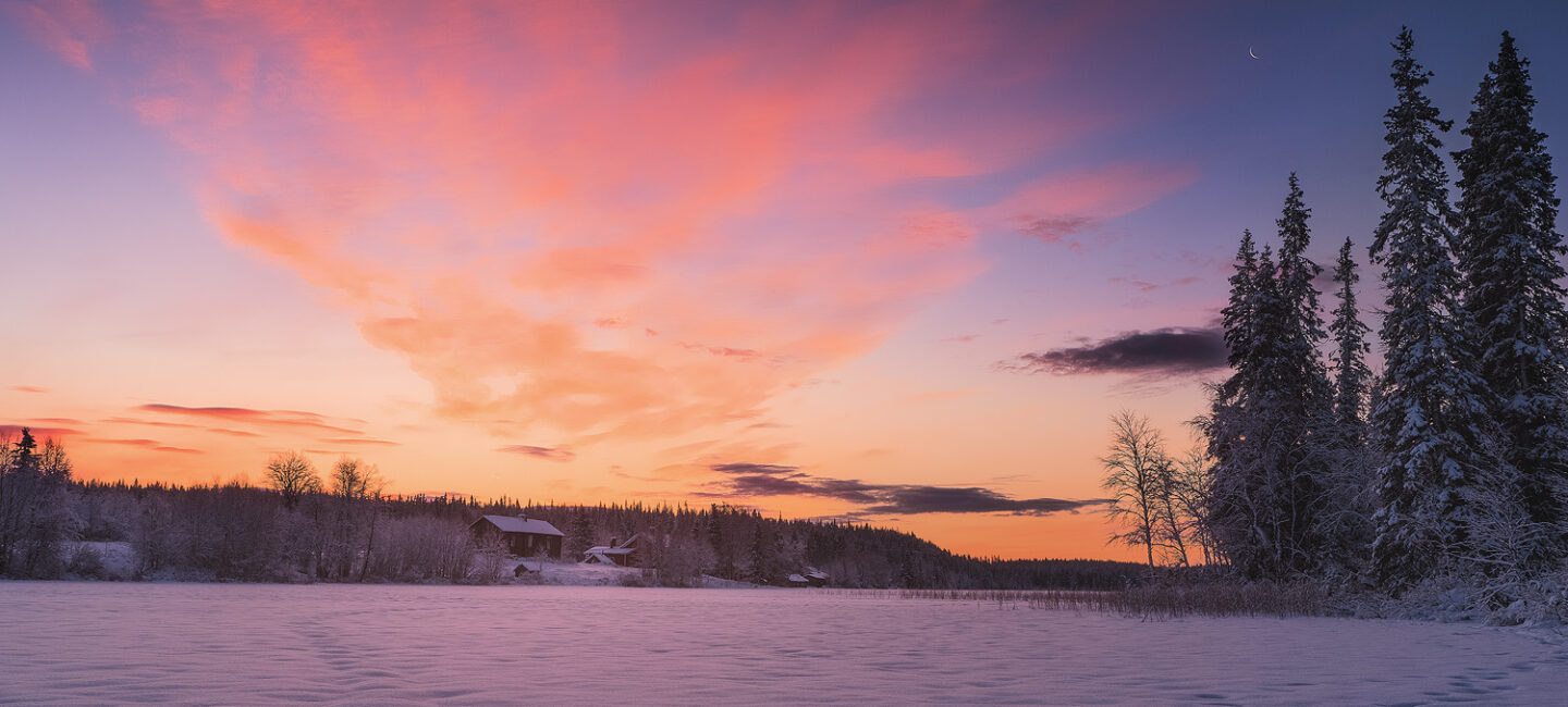 Sun rises above a small cabin along a frozen lake in Finnish Lapland