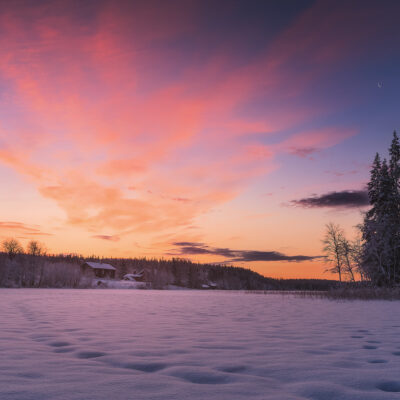 Sun rises above a small cabin along a frozen lake in Finnish Lapland